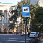 Placa azul de alerta para câmera de trânsito em uma rua urbana, com edifícios desfocados ao fundo