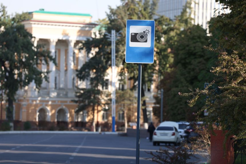 Placa azul de alerta para câmera de trânsito em uma rua urbana, com edifícios desfocados ao fundo