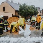 Equipe de emergência instala barreiras de sacos de areia em uma rua durante chuva, como medida de prevenção contra alagamentos.