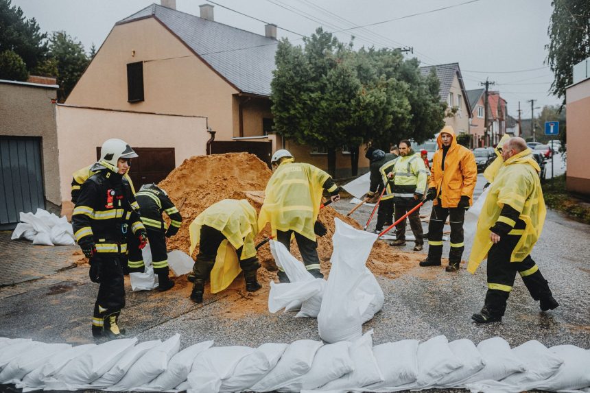 Equipe de emergência instala barreiras de sacos de areia em uma rua durante chuva, como medida de prevenção contra alagamentos.
