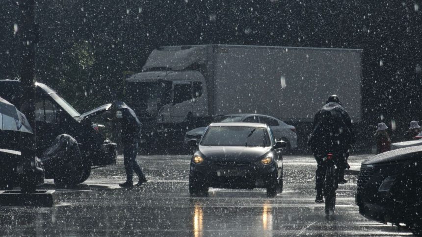Rua urbana com carros e pedestres sob chuva intensa.
