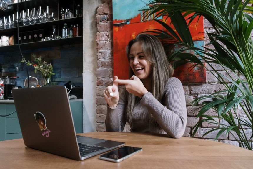 Mulher faz sinais em Libras durante uma videochamada em um laptop, em ambiente interno.