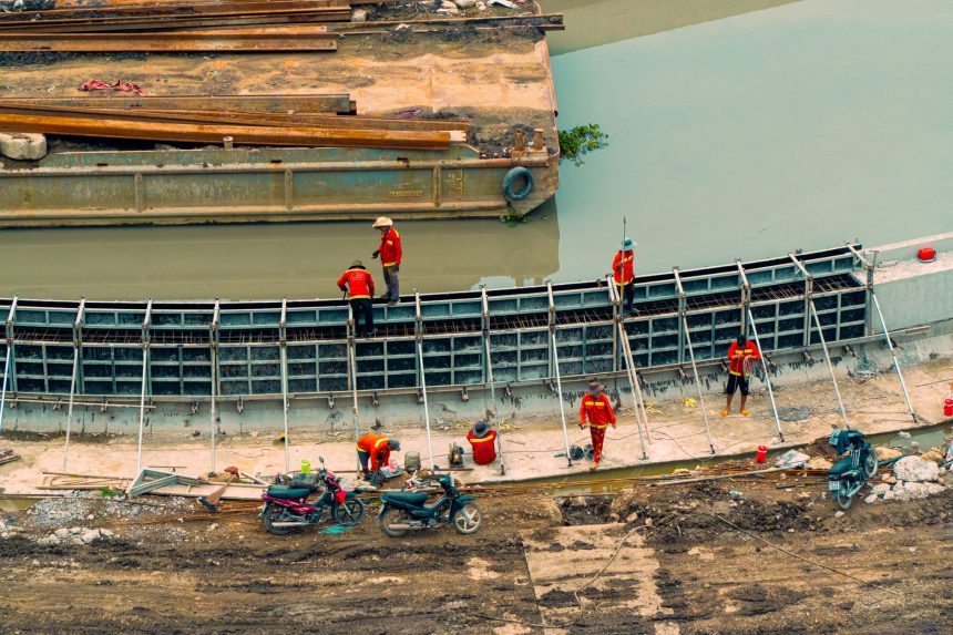 Vista aérea de trabalhadores e máquinas construindo uma estrutura de canalização junto a um curso d’água.