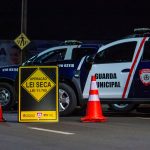 Agentes municipais realizam uma blitz de trânsito à noite, com viaturas e cones na via.