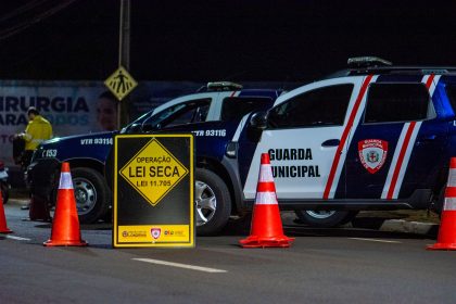 Agentes municipais realizam uma blitz de trânsito à noite, com viaturas e cones na via.
