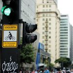 Semáforo e placa de travessia para bicicletas em uma rua movimentada de São Paulo.