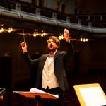 Maestro rege uma orquestra durante apresentação em uma elegante sala de concertos.